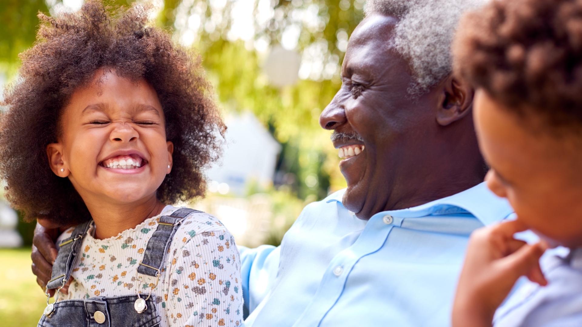Close-up of a Black grandparent and grandchild smiling and laughing outside with foliage blurred behind them.